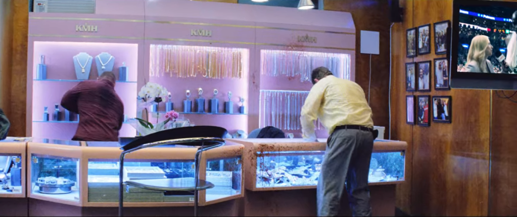 The store's main display counter is shown, with Phil in front bending over and and Nico at the display along the wall. Their backs are to us and they're both bent over. Blood spatter is visible on the display case to Phil's left, the store TV in the upper right corner of our view. The TV is cut seemingly in half, and the left half visible to us displays a blonde reporter holding a microphone and talking to KG, but he is cut in half also by the framing of the TV and overall image. Faint glare is visible below the TV, suggesting we see this scene from behind the security door glass where the gangsters just were.