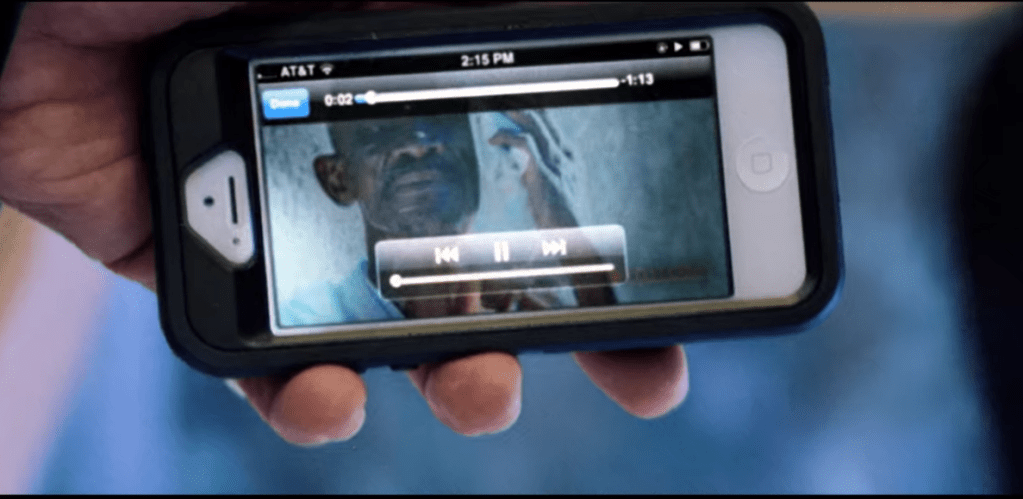 A close-up of Howard's hand holding a cell phone oriented horizontally. Video player controls are visible and on its screen is a black man with a mustache against a gray/black shadowy background, but other details are difficult to discern.