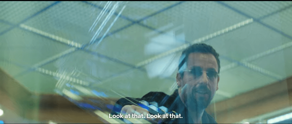 Ratner is shown from the lower perpsective behind the security glass, this time close enough that his face and upper torso are all that are visible. Multiple reflections of light create glare and other distortions. Behind Ratner the ceiling is visible, with square patterns formed by the ceiling structural supports.