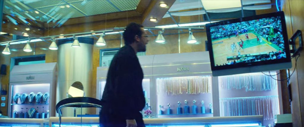 The interior of the jewelry store shown from a low perspective behind the security glass where Arno and gang are trapped. Ratner is centered and faces the TV screen in the upper right of the view. Behind Ratner are numerous glittering displays of watches, chains, and other items. Display lights create a line along the top quarter of the image and run between Howard and the television in the field of depth. The green of the basketball court is centered on that TV and seems to be the most distinctive detail