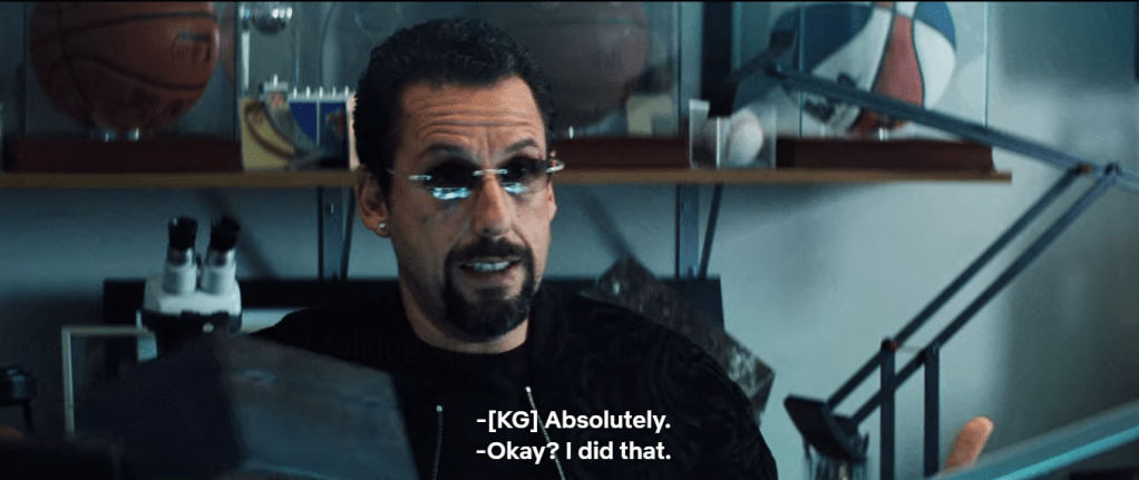 Howard is seated with basketball memorabilia on a shelf behind him, an orange basketball displayed to his back left and slight-right, a red, white, and blue baskbetball at the end of the shelf at the back right. A microscope is visible behind and to the left below the basketball shelf. He is wearing black glasses and black shirt. Subtitle: "-[KG] Absolutely. -Okay? I did that."