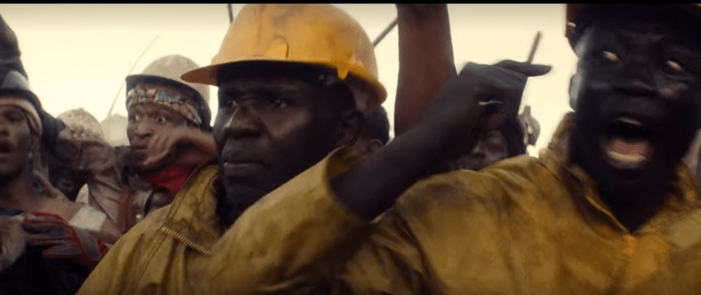 A black man with yellow protective jacket appears closest to the camera at the right. His mouth is open, eyes fixed on someone to the viewer's left but out of frame. He points at his eyes. To his left is another black man, mouth closed, in yellow gear and behind them is a crowd of other people, all focused in the same direction left of frame as the primary miner.