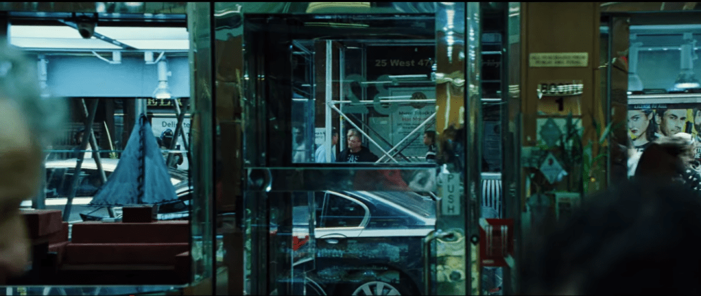 The camera shows a perspective from near where Howard has just been standing of the glass storefront and door. The glass is interspersed with brown/gold structures framing the door and other aspects of storefront. Poles and an apparent scaffolding appear on the other side of the glass and doors, creating numerous criss-cross and framing patterns. In the distance in the center of the shot, in the center of the door and framed in a triangle formed by scaffolding across the street is Nico, barely visible, and Phil, mostly obscured by a construction pole also framed within the door. People who could be diegetically outside or frozen images from a screen or poster crowd in a transparent section of storefront on the viewer's right. Fragmented reflections caught in glass are strewn throughout the image.