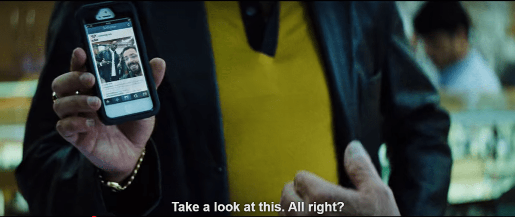 Howard Ratner's torso takes up the majority of the image, wearing a yellow/gold shirt beneath black jacket. His right hand is holding up his phone, on which appears a small image of him and KG taken earlier in the jewelry store. Subtitle: "Take a look at this. All right?"