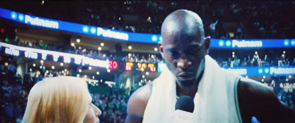 Kevin Garnett with a white towel around his shoulders, on top of white and green Celtics shirt, speaking with woman reporter holding a microphone on the floor of the crowded auditorium. The in-movie TV image of Garnett takes up the full screen.